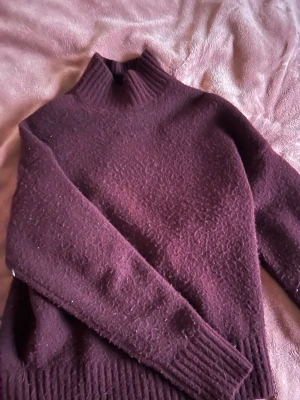 Vinröd stickad polotröja - Mysig vinröd stickad polotröja med hög ribbad krage och breda ribbade muddar vid ärmslut och nederkant. Tröjan har en rak passform och är tillverkad i ett mjukt, tjockt material som passar perfekt för kyliga dagar.