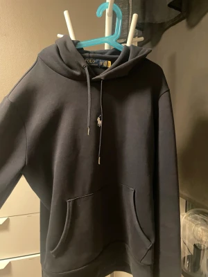Mörkblå hoodie från Polo Ralph Lauren - Mörkblå hoodie från Polo Ralph Lauren med klassisk huva och dragsko. Har en stor magficka och den ikoniska vita Polo-loggan broderad på bröstet. Väldigt bra skick, pris kan diskuteras och hör av dig vid minsta fundering! 