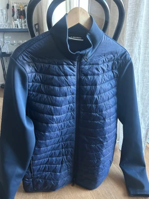 Marinblå jacka från J.Lindeberg - Snygg marinblå vindjacka från J.Lindeberg med quiltad front och ryggparti. Jackan har släta ärmar, hög krage och dragkedja framtill. Materialet är en mix av glansigt och matt tyg som ger en modern look. Perfekt för blåsiga dagar. Köptes i vintras. Inga skador som ny.