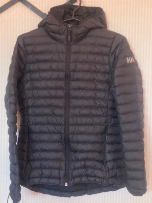Svart dunjacka med huva Helly Hansen - Snygg svart dunjacka från Helly Hansen med huva och dragkedja framtill. Jackan har quiltad design, två sidofickor med dragkedja och diskret HH-logga på ärmen. Perfekt för kalla vinterdagar och riktigt lätt att bära.