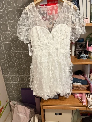 Vit studentsklänning med blommor - Superfin vit kort klänning med v-ringning och transparenta spetsärmar. Klänningen har 3D-blommor över hela tyget och volangkant nedtill. Perfekt för dig som vill ha en romantisk och unik look. Puffiga korta ärmar och omlottkänsla vid bysten.
