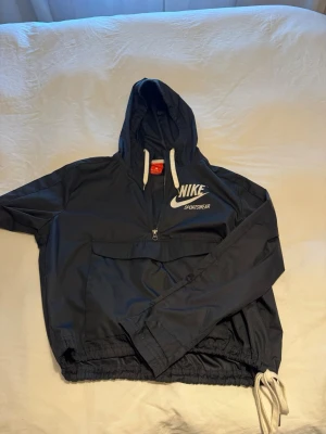 Svart Nike vindjacka med huva - Snygg mörkblå vindjacka från Nike med stor vit logga och texten 'Sportswear' både fram och bak. Jackan har huva med vita dragskor, halv dragkedja framtill och en stor magficka. Resår i midjan och vid ärmslut för skön passform. Perfekt för en sportig look.