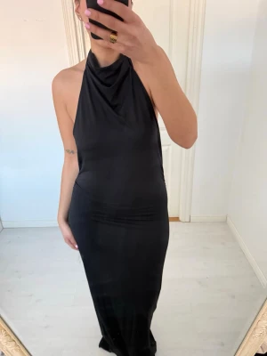 Svart lång halterneckklänning - Stilren svart långklänning med halterneck och figurnära passform. Klänningen är ärmlös och har en mjuk, glansig yta som ger en elegant look. Perfekt för dig som vill ha en enkel men snygg outfit.