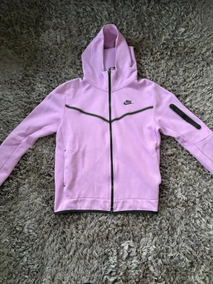 Ljuslila Niketech med dragkedja - Nike hoodie i ljuslila med hel dragkedja och svart Nike-logga på bröstet. Snygg detalj med svart linje över bröstet och ficka med dragkedja på ena ärmen. Mjuk bomullskänsla och huva för extra stilpoäng.