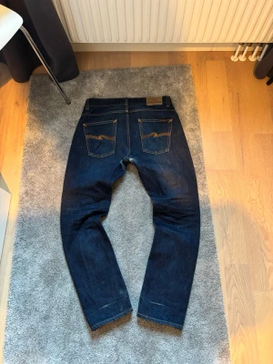 Nudie jeans - Säljer ett par mörkblå jeans från Nudie Jeansen har kontrastsömmar och Nudie-logga på bakfickorna. Tillverkade i kraftig denim, perfekta för en stilren vardagslook. Dem har en rak passform och fått sköna fades samt slitningar efter en dels bärande. 