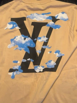 Beige L…..V t-shirt med molntryck - Snygg beige t-shirt  med stort L..Vtryck täckt av blåvita moln på ryggen och mindre tryck framtill. Klassisk passform och rund hals. Perfekt för dig som vill sticka ut med en unik design.