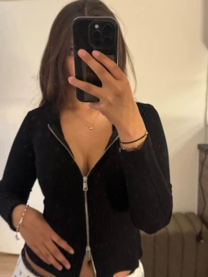 Svart ribbad ziptröja - Svart ribbad tröja med dragkedja framtill och djup v-ringning. Långa ärmar och figurnära passform, perfekt för en trendig och avslappnad look.