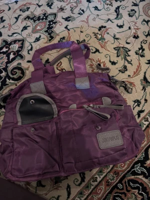 Lila handväska från JINGPINWOLF - Cool lila handväska i nylon med flera fickor framtill och grå detaljer. Väskan har dubbla handtag och dragkedjor, samt en logotyp på framsidan. Perfekt för dig som gillar praktiska och snygga accessoarer.