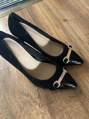 Svarta pumps från Anna Field - Snygga svarta pumps från Anna Field med spetsig tå och dekorativt guldfärgat spänne framtill. Skorna har en mix av matt och blank finish, där tån är i lack och resten i mocka. Perfekt för dig som gillar stilrena detaljer och klassisk design.