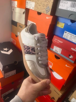 Sneaker med leopardmönster och metallic - Coola sneakers från Tamaris med vit bas, metallic-silver detaljer och en leopardmönstrad panel på sidan. Skon har klassisk låg profil, vita skosnören och en ljusbrun gummisula. Perfekt för dig som vill sticka ut med din stil.