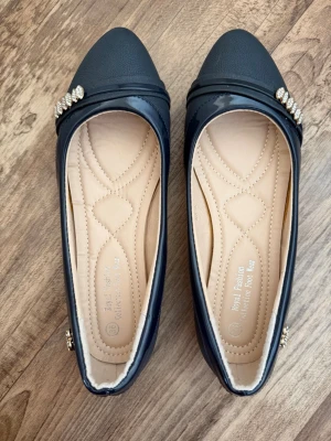 Mörkblåa ballerinaskor - Snygga mörkblåa ballerinaskor från Royal Fashion med spetsig tå och dekorativa strassdetaljer på ovansidan. Skorna har en blank finish och mjuk, quiltad innersula i beige för extra komfort. Perfekt för dig som vill ha en elegant och stilren look. 