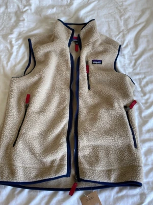 Beige fleeceväst från Patagonia - Snygg beige fleeceväst från Patagonia med blå detaljer och röda dragkedjor. Västen har hög krage, tre fickor med dragkedja och är tillverkad i mjukt, värmande fleece-material. Perfekt för lager-på-lager och outdoor-vibe.