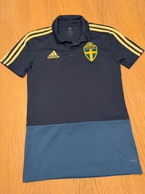 Blå sverige landslags tröja  - Mörkblå pikétröja från Adidas med Sveriges landslagsemblem på bröstet och gula detaljer. Tröjan har klassisk krage, knappar framtill och gula ränder på axlarna. Tillverkad i funktionsmaterialet Climalite, perfekt för träning eller support.