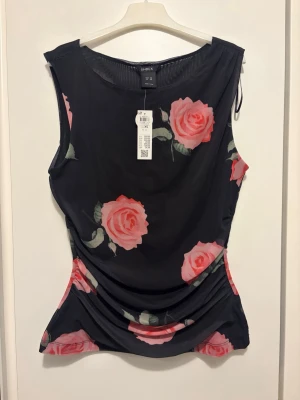 Svart blommig ärmlös topp Lindex - Snygg svart ärmlös topp från Lindex med stora rosa rosor som mönster. Toppen har en rund halsringning och lätt rynkade sidor för en smickrande passform. Tillverkad i ett mjukt och stretchigt syntetmaterial. Ordinarie pris 299kr.