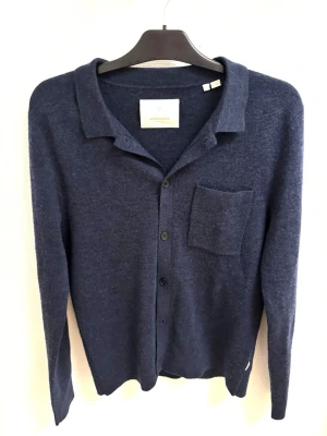 Mörkblå kofta från Jack & Jones  - Stilren mörkblå kofta från ARKET med knäppning framtill och en bröstficka. Koftan har klassisk krage och är tillverkad i ett mjukt, stickat material. Perfekt för lager-på-lager och enkel att matcha med olika outfits.