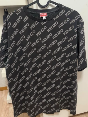 Grå Kenzo t-shirt med logotypmönster - Snygg svart t-shirt från Kenzo med grått logotypmönster över hela plagget. Klassisk rund halsringning och korta ärmar. Perfekt för dig som gillar streetwear och vill sticka ut med ett trendigt designerplagg. Skriv om ni behöver fler bilder ❤️