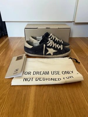 Svarta Golden Goose sneakers med stjärna - Säljer ett par svarta Golden Goose sneakers med vit stjärna på sidan och vita detaljer. Skorna har vita snören och en vit häl, samt en vintage-inspirerad look. Materialet är en mix av läder och textil. Medföljer originalkartong och dustbag.