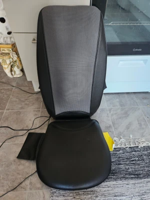 Svart massagekudde för stol (oanvänt) - Elektrisk massagekudde i svart konstläder och grått tyg, designad för att placeras på en stol. Har sladd för ström och passar perfekt för rygg och säte. Enkel att använda hemma för avslappning.                                                        (Nytt oanvänt)