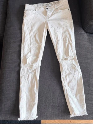 Vita super skinny jeans med slitningar - Säljer ett par vita super skinny jeans med ankellängd och slitna detaljer vid knäna. Jeansen har råa kanter vid bensluten och klassisk femficksdesign. Perfekta för dig som gillar en tight och trendig look.