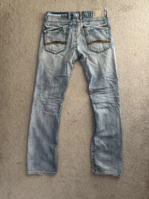 Ljusblå jeans från SMOG med raka ben - Ljusblå jeans från SMOG med raka ben och slitna detaljer. Jeansen har fem fickor, synliga knappar i gylfen och kontrastsömmar. Bakfickorna har en unik design med snedställda sömmar och dekorativa detaljer. Perfekta för en avslappnad streetstil. Har några defekter som en fläck som knappt syns och ett hål på låret.