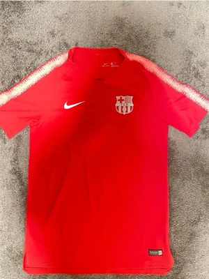 Röd FC Barcelona träningströja Nike - Röd träningströja från Nike med FC Barcelona-logga på bröstet och vita detaljer längs axlarna. Tröjan har korta ärmar och är tillverkad i ett lätt och ventilerande material, perfekt för fotbollsträning eller sportaktiviteter.