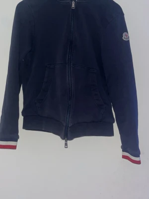 Mörkblå hoodie från Moncler - Snygg mörkblå hoodie från Moncler med dragkedja framtill och fickor på magen. Ärmsluten och huvan har rödvita ränder som ger en sportig vibe. Moncler-logga på ärmen. Perfekt för dig som gillar streetwear och exklusiva märken.