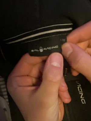 Svart fleecejacka från Sail Racing - Svart fleecejacka från Sail Racing med hel dragkedja och logotyp på bröstet. Jackan har ribbade detaljer vid kragen och är tillverkad i mjukt fleecematerial. Perfekt för lager-på-lager och håller dig varm under kyliga dagar.