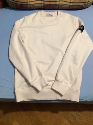 Vit sweatshirt från Stone Island - Säljer en stilren vit sweatshirt från Stone Island med klassisk rund hals och ribbade muddar. Tröjan har den ikoniska Stone Island-loggan på vänster ärm och är tillverkad i mjukt bomullsmaterial. Perfekt för dig som gillar minimalistisk design.