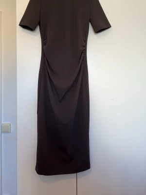 Mörkbrun långklänning med drapering - Säljer en stilren mörkbrun långklänning med korta ärmar och snygg drapering i midjan. Klänningen har en rak passform och är tillverkad i ett mjukt, stretchigt material som ger en bekväm känsla. Perfekt för dig som gillar minimalistisk och elegant stil.