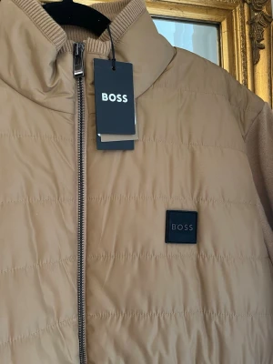 Beige pufferjacka från BOSS - Stilren beige pufferjacka från BOSS med quiltad design och svart logotyp på bröstet. Jackan har hög krage, dragkedja framtill och ribbade detaljer vid halsen. Helt ny med prislapp kvar, storlek M.. Priset går att diskutera men accepterar inte för låga priser