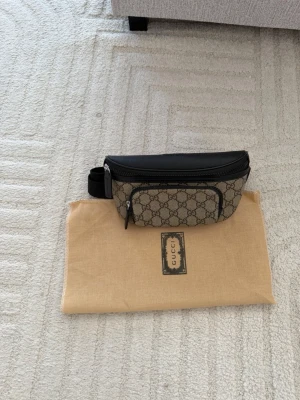 Gucci axelväska med GG-mönster - Snygg axelväska från Gucci i beige och svart med klassiskt GG-mönster. Väskan har svarta läderdetaljer, justerbar rem och dragkedja framtill. Perfekt för dig som vill ha en trendig och praktisk accessoar med lyxig känsla.