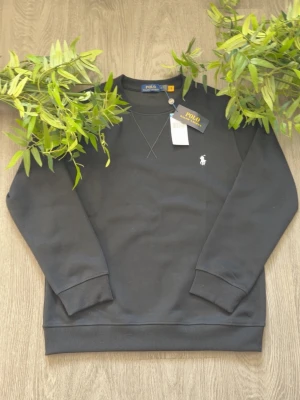 Svart sweatshirt från Polo Ralph Lauren - Svart sweatshirt från Polo Ralph Lauren med klassisk rund hals och broderad vit logga på bröstet. Tröjan har långa ärmar och ribbade muddar vid ärmslut och nederkant. Perfekt för en stilren och avslappnad look. Kvittot finns vid behov och fråga gärna om bilder eller info