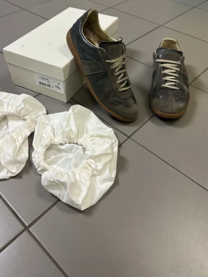 Maison Margiela gats - Maison Margiela sneakers i grå mocka och skinn med vit snörning och gummisula. Klassisk low-top siluett med rund tå och diskret branding på sulan. Tillverkade i Italien, stilrena och passar perfekt till streetwear eller casual outfits sista bilderna är tydliga och du som köper ser hur de ser utt. Mitt pris 2399kr !ej hugget i sten 🪨!