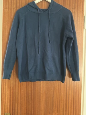 Blå merinoull hoodie med huva - En enkel och stilren blå hoodie med huva och dragsko. Hoodien har långa ärmar och ribbade muddar vid ärmslut och nederkant. Perfekt för en avslappnad look och passar till det mesta. Materialet känns mjukt och bekvämt.