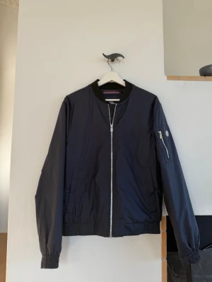 Mörkblå bomberjacka från Snoot - Snygg mörkblå bomberjacka från Snoot med silverfärgad dragkedja, ribbade muddar och svart krage. Jackan har en ficka med dragkedja på ärmen och två sidofickor. Tillverkad i ett lätt, glansigt material som ger en modern look. Passar 1.80 cm cirka. 