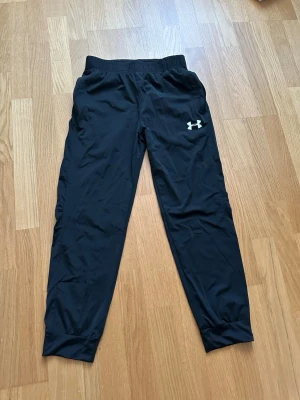 Svarta träningsbyxor Under Armour - Säljer ett par svarta träningsbyxor från Under Armour med resår i midjan och smal passform. Byxorna har logga på låret och texten UNDER ARMOUR längs sidan. Perfekta för gymmet eller löpning, tillverkade i ett lätt och snabbtorkande material.