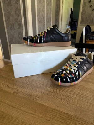 Maison Margiela med färgstänk - Unika svarta sneakers från Maison Margiela med färgstänk i flera färger över hela ovandelen. Skorna har beige sula och vita snören. Tillverkade i läder med låg profil och klassisk siluett. Perfekta för dig som vill sticka ut med din stil.