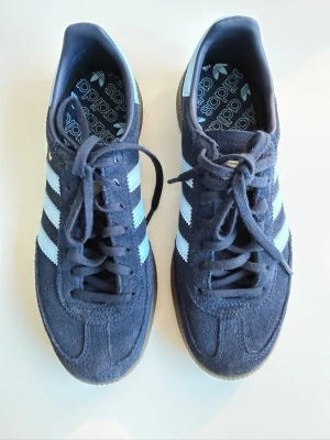 Adidas Gazelle blå sneakers - Säljer ett par klassiska Adidas Gazelle sneakers i mörkblå mocka med vita ikoniska stripes på sidorna och gummisula. Skorna har låg profil och snörning framtill. Perfekt för dig som vill ha en stilren och tidlös look.