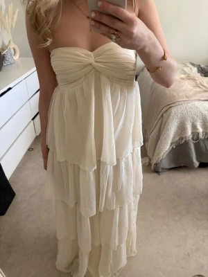 Ljus axelbandslös långklänning - Romantisk ljus långklänning med flera lager volanger och twistad detalj vid bysten. Axelbandslös modell som ger en elegant siluett. Perfekt för sommarens alla tillfällen. Materialet känns lätt och luftigt.