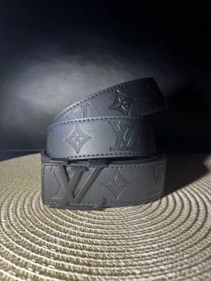 Svart skärp  - Snyggt svart skärp med präglad monogramdesign och spänne. Bältet är gjort i skinn och har en stilren look som passar till många outfits. Perfekt accessoar för dig som gillar exklusiva detaljer.