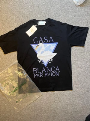 Svart Casablanca t-shirt med svanmotiv - Svart t-shirt från Casablanca med stort tryck av en vit svan och texten 'Casa Blanca Par Avion' i lila framtill. Klassisk passform och rund hals. Tillverkad i mjuk bomull, perfekt för en avslappnad och trendig look.
