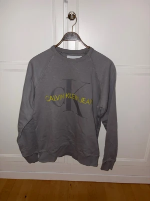 Grå sweatshirt Calvin Klein - Snygg grå sweatshirt från Calvin Klein Jeans med stort CK-tryck och gul text på bröstet. Tröjan har rund halsringning, långa ärmar och ribbade muddar vid ärmslut och nederkant. Perfekt för en avslappnad och trendig look.