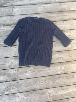 Mörkblå kabelstickad tröja Polo Ralph Lauren - Kabelstickad mörkblå tröja från Polo Ralph Lauren med rund halsringning och ribbade muddar. Tröjan har det klassiska röda Polo-logot broderat på bröstet och är tillverkad i mjukt material. Perfekt för en stilren och avslappnad look.