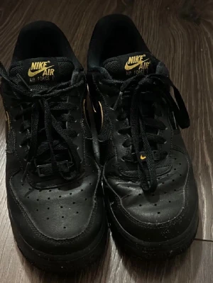 Nike Air Force 1 svart/guld sneakers - Säljer ett par Nike Air Force 1 sneakers i svart med gulddetaljer på loggan och snörningen. Skorna har klassisk låg profil, perforerad tå och är tillverkade i läder med textildetaljer. Väl använda.