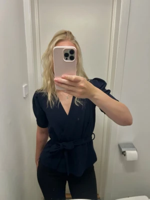 Mörkblå blus - Snygg mörkblå omlottblus från Sisters Point med knytband i midjan och v-ringning. Blusen har korta puffärmar och är tillverkad i ett mjukt, följsamt tyg som ger en elegant siluett. Perfekt att styla med både jeans och kjol.