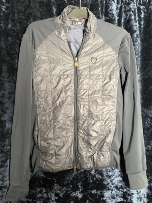 Grå vindjacka från EA7 Emporio Armani - Snygg grå vindjacka från EA7 Emporio Armani med quiltad framsida och släta ärmar. Jackan har dragkedja framtill, hög krage och ett EA7-märke på bröstet. Materialet är en mix av glansig nylon och stretchigt tyg på ärmarna. Perfekt för en sportig och stilren look. 