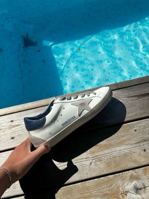 Golden goose skor - Snygga vita sneakers med grå stjärndetalj på sidan och mörkblå häl. Skorna har grå snören och en klassisk låg siluett. Materialet ser ut att vara läder med mockadetaljer. Perfekta för en trendig och avslappnad stil. Innersulan är 23 och en halv😁
