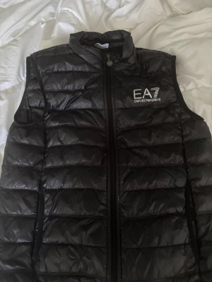 Svart dunväst från EA7 Emporio Armani - Snygg svart dunväst från EA7 Emporio Armani med quiltad design och hög krage. Västen har dragkedja framtill och logga på bröstet. Perfekt för lager-på-lager och ger en sportig vibe. Tillverkad i glansigt material som känns modernt och trendigt. ( det står att den är storlek L men hade sagt att den är mer åt M hållet.)