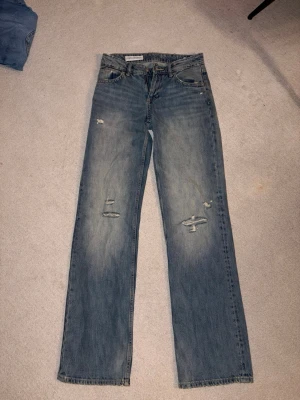 Blå slitna raka jeans - Säljer ett par blå jeans från Zara med rak/bage passform och slitna detaljer både fram och bak. Jeansen har klassisk femficksdesign och tydliga slitningar på låren och baksidan av ena benet. Perfekta, trendig look.