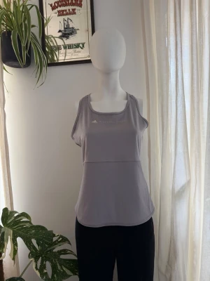 Adidas Stella McCartney 2017 Ljusgrå träningstop - Tränings topp från Adidas Stella McCartney med sportig design och rundad halsringning. Tillverkad i ett mjukt och stretchigt syntetmaterial som passar perfekt för träning. Diskret logga framtill. Kollektionen 2017, finns några små imperfections se bilder. 